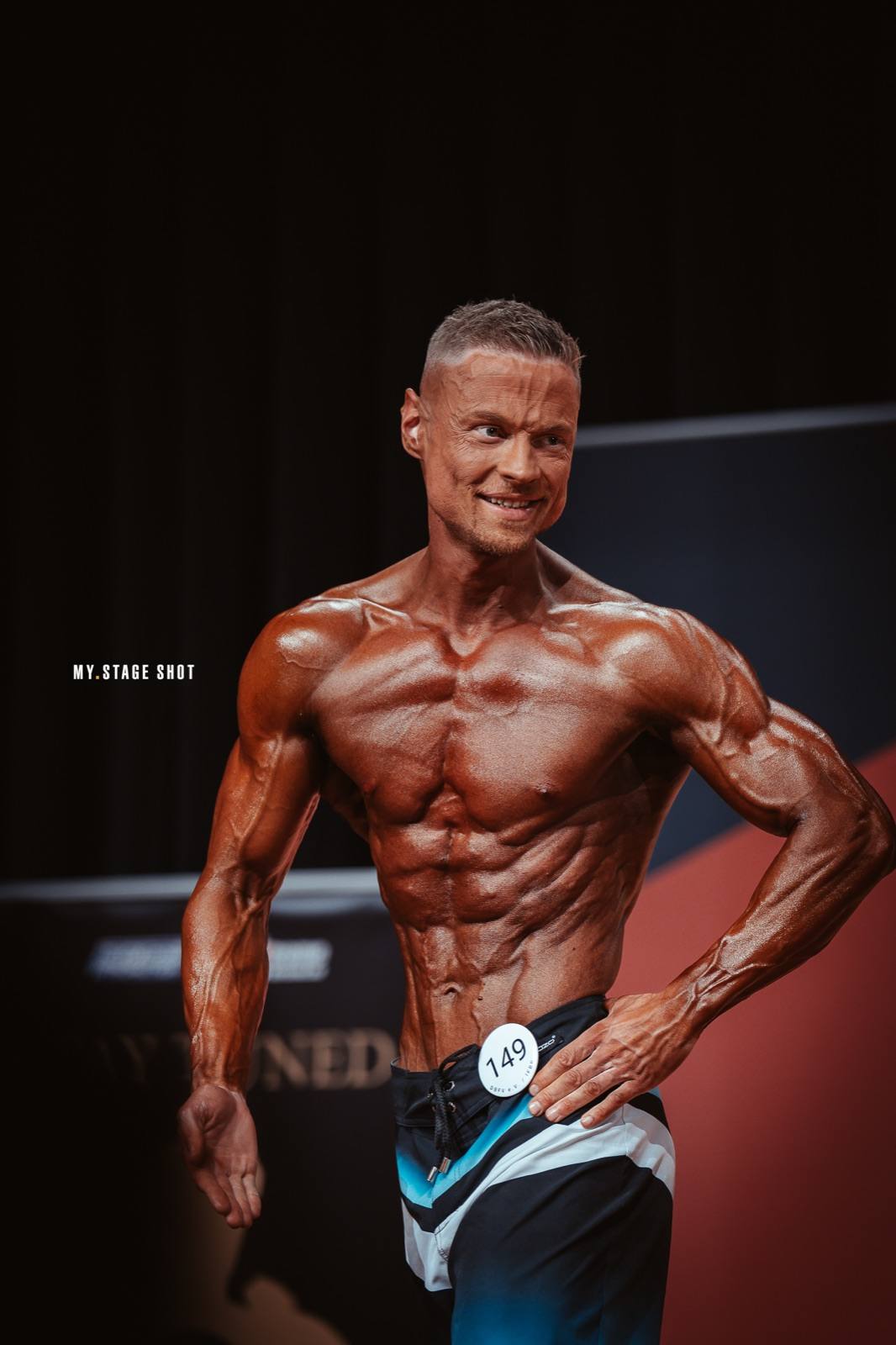 Men’s-Physique-Athlet in der Seitpose vor dunklem Vorhang